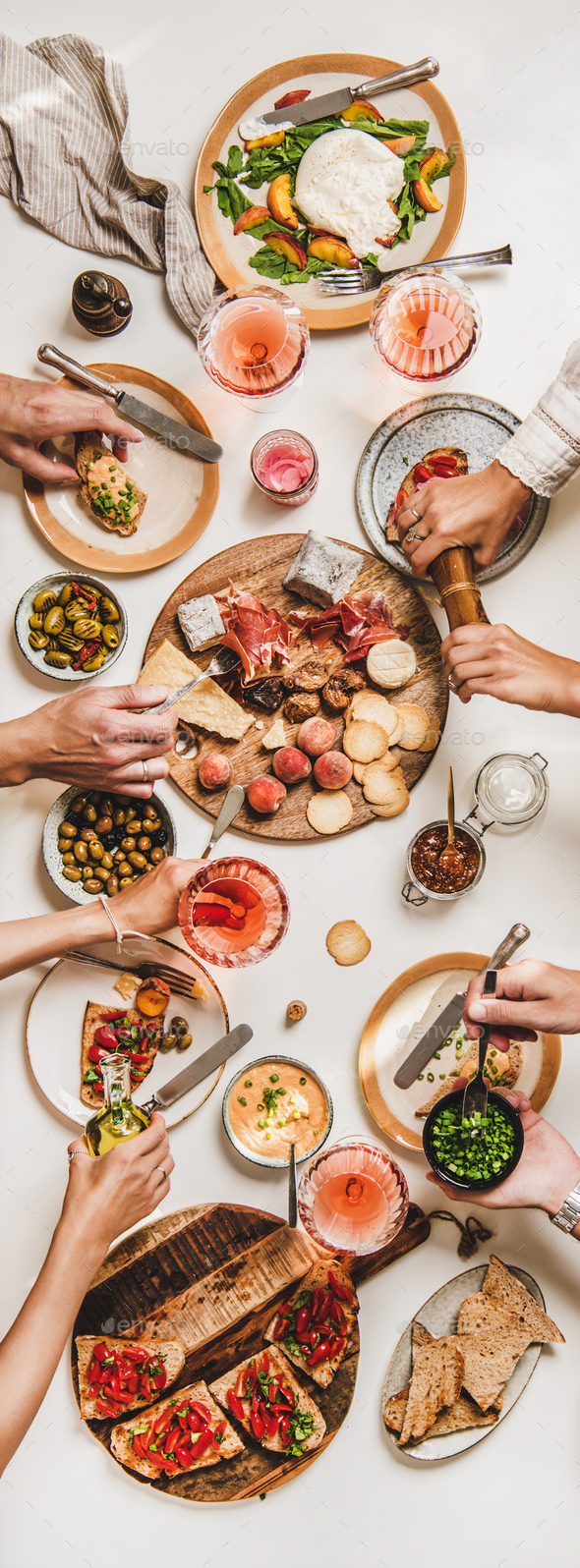 Family wine and snack party. Flay-lay of peoples hands eating and drinking rose wine over table with cheese, fruit, smoked meat, tomato brushettas, buratta salad, top view. Wine tasting concept
