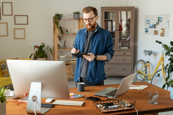Young creative software developer or designer drawing on tablet display in front of computer monitor while standing in by his workplace at home