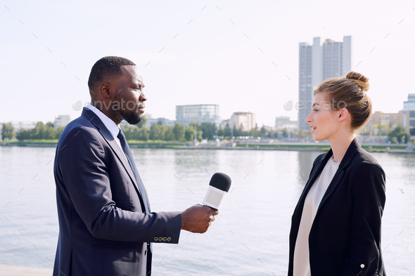 Young male reporter of African ethnicity standing in front of successful businesswoman while taking interview in urban environment