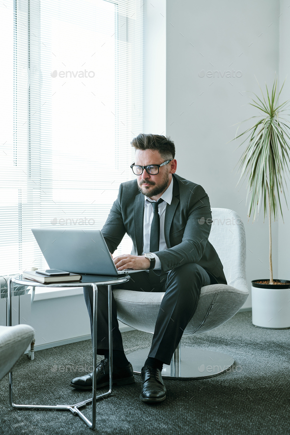 Young elegant businessman in formalwear using laptop while sitting in soft comfortable armchair by small table against large window in office
