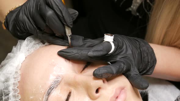 Beautician in Black Gloves Does Permanent Makeup Correction of the Shape of the Eyebrows to Young