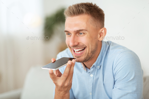 Voice Search Concept. Closeup portrait of young bearded guy using voice assistant or calling on smartphone at home, blurred background, copy space. Smiling man talking on mobile phone