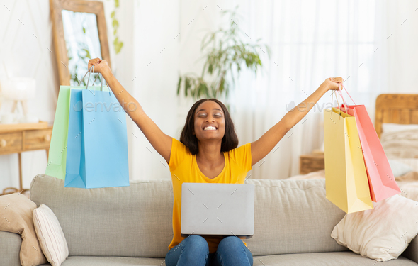 Excited black lady with laptop lifting hands full of shopping bags on sofa at home. Lovely African American woman feeling happy over big sale in online shop, purchasing goods over internet indoors