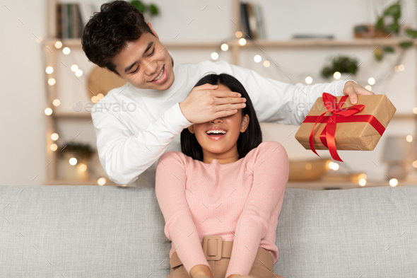 New Year Celebration. Asian Husband Giving Christmas Gift To Wife, Covering Her Eyes Celebrating Holiday Sitting On Coush At Home. Birthday And Xmas Presents, Happy Winter Holidays Concept