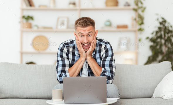 Great News Concept. Portrait of emotional guy celebrating online victory sitting on the sofa at home, touching his face, using laptop computer, copy space. Shocked male model looking at pc