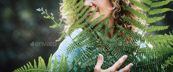 Concept of earth’s day love and celebration with closeup portrait of smiling adult woman hugging a big tree leaf in outdoor nature forest – green defocused background
