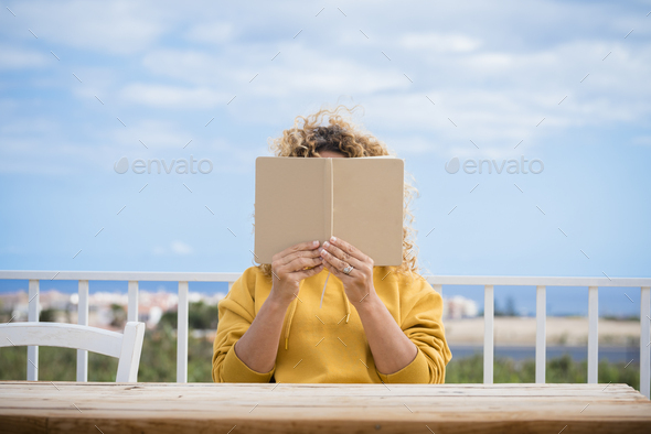 Unrecognizable woman read a paper book outdoor with wooden table in front of her and blue beautiful sky in background – concept of reading and studying for people