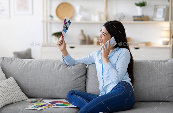 Cheerful Korean Woman Choosing Design Of New Apartment, Holding Color Palette And Talking On Cellphone At Home, Discussing Project With Designer, Sitting On Couch In Living Room, Copy Space