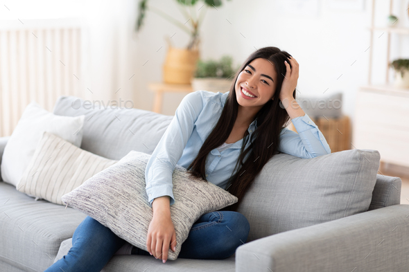 Domestic Portrait. Cheerful Korean Woman Posing On Comfy Couch At Home, Cuddling Pillow, Smiling And Looking At Camera, Resting In Cozy Light Living Room, Enjoying Relaxed Weekend, Copy Space