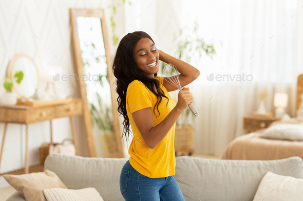 Joyful African American woman using whisk as microphone to sing and dance at home. Millennial black lady pretending to be rockstar, having great time in living room