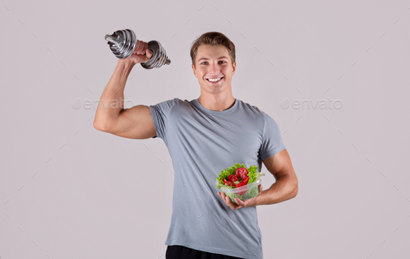 Healthy diet and exercising. Happy Caucasian guy lifting dumbbell and holding vegetable salad on