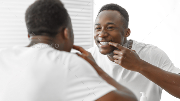 Toothcare Concept. African American Man Checking Perfect White Teeth Smiling To His Reflection In Mirror Standing In Bathroom At Home. Teeth Health And Everyday Oral Hygiene Concept. Panorama