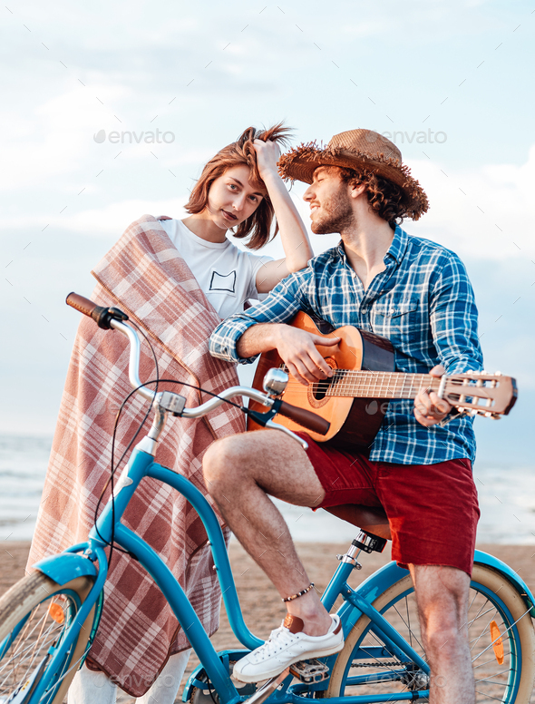 Two young people with their bicycle. Boy is playing on the guitar. Girl is wrapped in pink blanket, her arm is on the boy’s shoulder