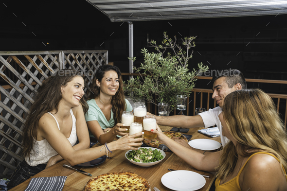 Friends Cheering With Beer Glasses Sitting Around a Table With Pizzas and Salads. - Stock Photo - Images