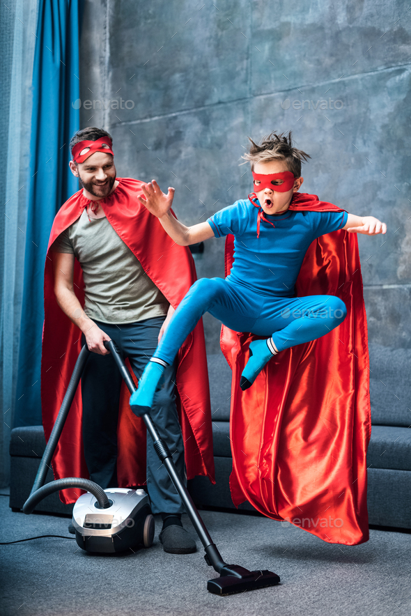 father in superhero costume vacuuming carpet while son in superhero costume jumping