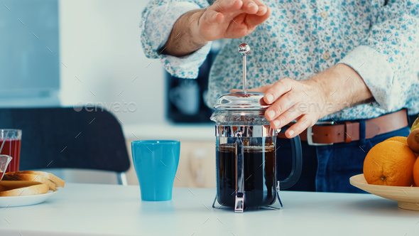 Old man pushing down the lid of french press while making coffee for breakfast in kitchen. Elderly person in the morning enjoying fresh brown cafe espresso cup caffeine from vintage mug, filter relax refreshment