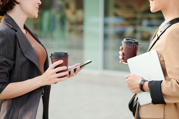 Two young successful contemporary businesswomen with drinks and mobile gadgets standing in front of each other against building exterior