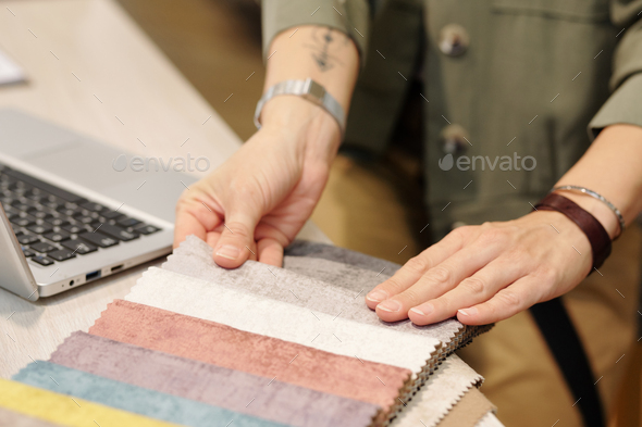 Hands of young female manager of studio of interior design looking through samples of new textile for furniture in front of laptop
