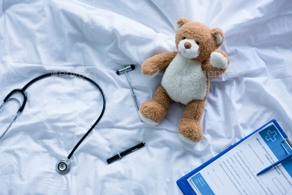 Stethoscope, reflex hammer and clipboard with medical document lying on bed near teddy bear