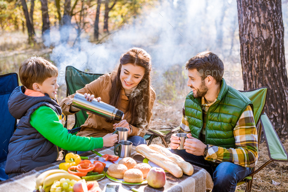 Happy smiling family drinking tea while sitting at table on picnic in autumn forest