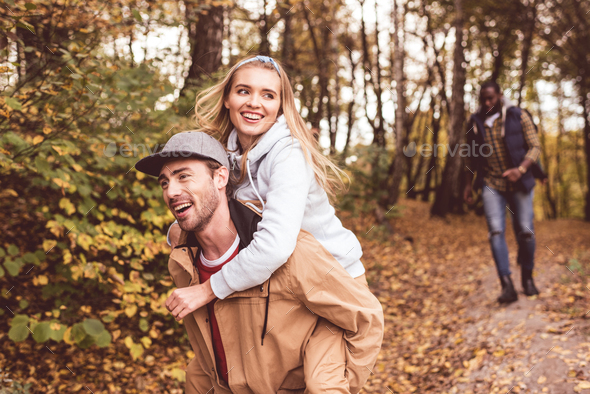 Happy young couple piggybacking and having fun in autumn forest with blurred running man