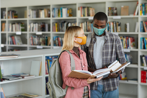Waist up portrait of two students wearing masks while standing in school library and holding books, copy space
