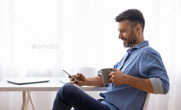 Break In Work. Relaxed Male Freelancer Using Smartphone And Drinking Coffee While Sitting At Desk At Home Office, Browsing Internet Or Social Networks, Enjoying Remote Job, Side View With Copy Space