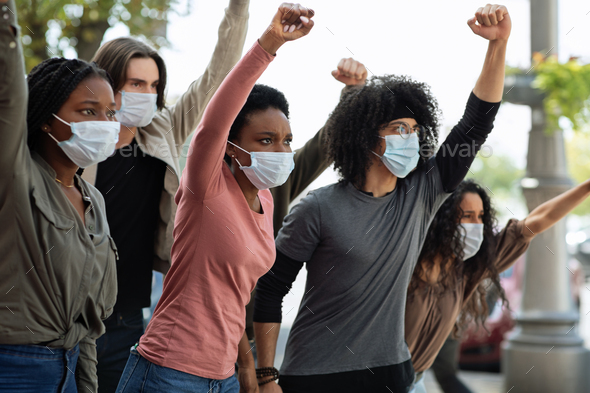 Group of indignant multiracial students protesting against racism on the street. Young people in face masks yelling slogans and raising clenched fists up, expressing disagreement with government