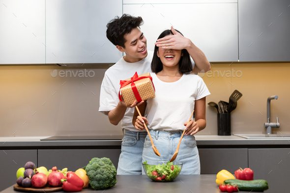 Family Holiday Celebration. Asian Husband Preparing Surprise Gift For Happy Wife Standing In Kitchen At Home. Couple Celebrating Birthday And Cooking Romantic Celebratory Dinner