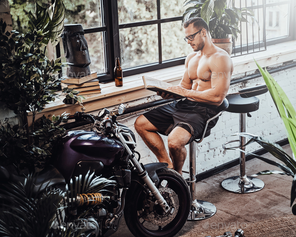 Strong athletic man with book sits on chair near motorcycle in exotic and unique hall room.