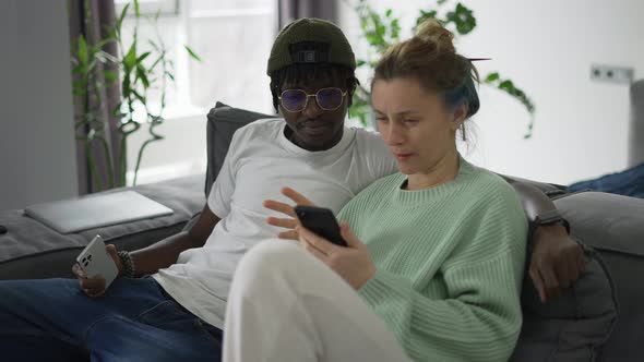Happy Biracial Couple Relaxing on Sofa with Smartphone alt