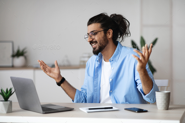 Excited indian man emotionally celebrating business success with laptop computer in office, sitting at desk and raising hands with joy, received good news, got promotion, finished project, free space