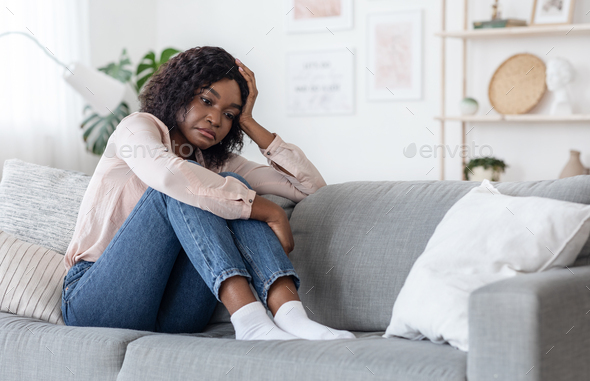 Seasonal Depression. Lonely Black Woman Sitting On Couch At Home With Sad Face Expression, African Lady Having Autumn Anxiety, Feeling Tired And Bored, Staying Alone In Light Living Room, Copy Space