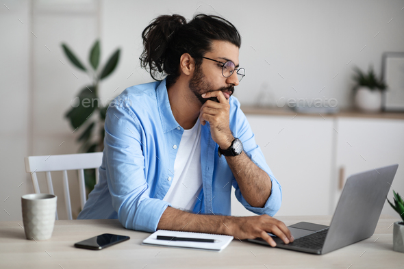 Thoughtful Indian Freelancer Working With Laptop At Home Office, Sitting At Desk Looking At Computer Screen, Thinking About Project Solution, Browsing Information In Internet, Copy Space