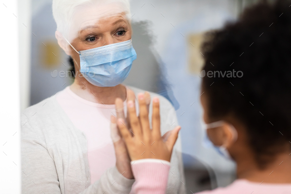 Coronavirus Sick Senior Female Patient Touching Grandchild’s Hand Through Glass Door Standing In Hospital. Covid-19 Pandemic, Corona Virus Self-Isolation Quarantine. Selective Focus