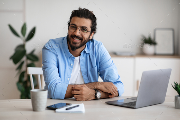 Freelance Career. Handsome Indian Guy In Casual Wear Smiling At Camera While Sitting At Laptop Desk In Home Office, Young Cheerful Western Man Enjoying Working Remotely Online On Computer, Copy Space