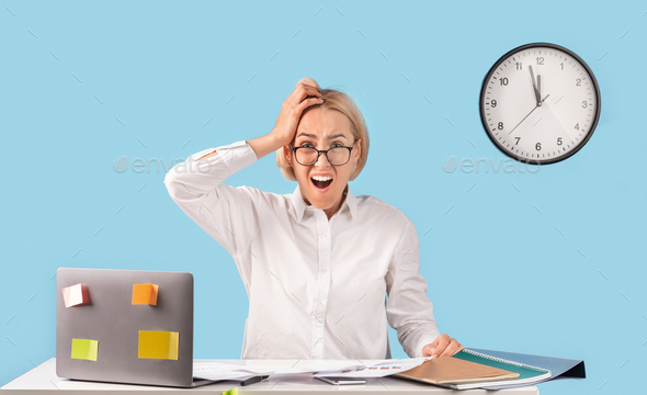 Caucasian business lady holding her head in terror, missing deadline at her desk over blue studio - Stock Photo - Images
