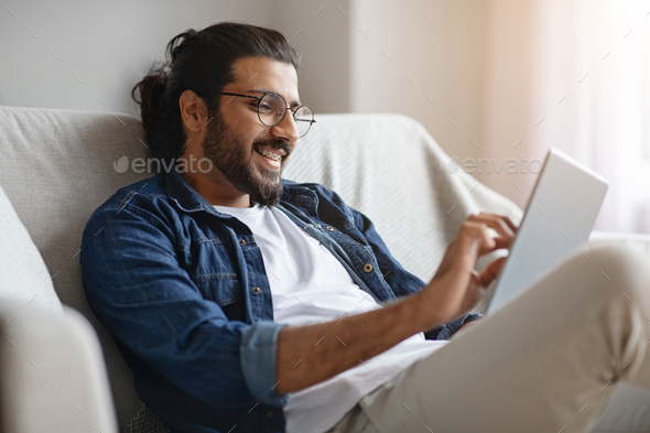Happy western man checking social media on digital tablet at home, sitting on comfortable couch in living room, browsing internet or new app, enjoying domestic passtime and modern technologies