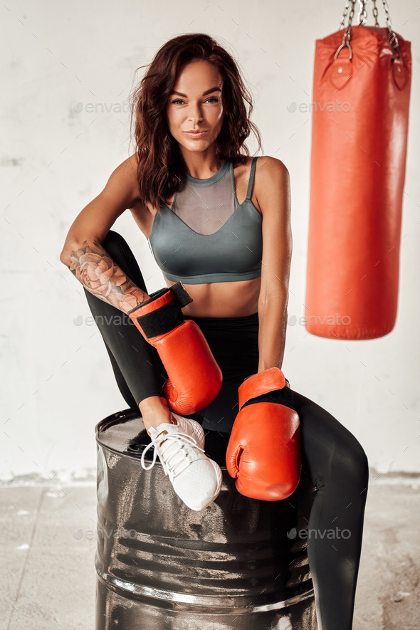 Beauty female boxer with red gloves resting after intense workout and sitting on barrel with punching bag on the background.