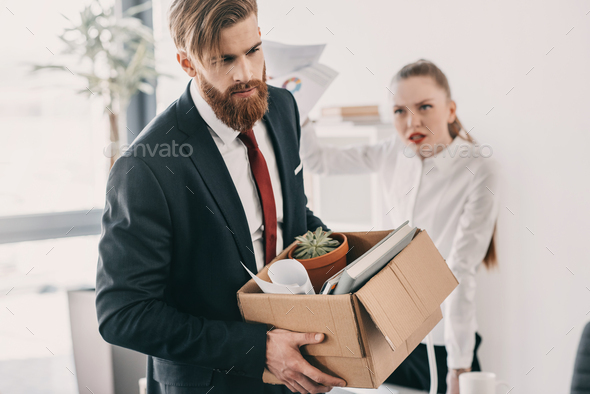 Young upset fired businessman with cardboard box and angry businesswoman in office - Stock Photo - Images
