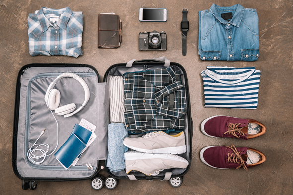 Top view of an essential vacation items in open luggage with digital devices on wooden background