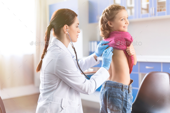 Young woman doctor with stethoscope listening to heartbeat and lungs of little girl patient
