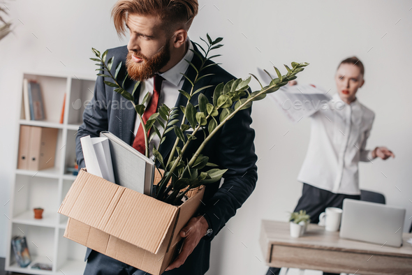 Young upset fired businessman with cardboard box in office, angry boss behind