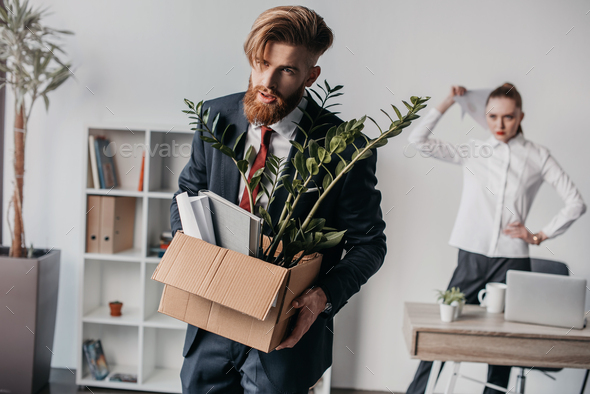 Young upset fired businessman with cardboard box in office, angry boss behind