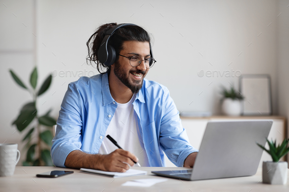 Distance Learning. Happy Indian Man In Wireless Headphones Watching Webinar On Laptop Computer And Taking Notes, Having Online Training Or Lecture, Sitting At Desk At Home Office, Free Space