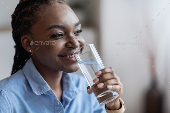 Stay Hydrated. Smiling African American Lady Drinking Mineral Water From Glass And Looking Away, Black Woman With Braids And Braces Enjoying Healthy Liquid, Closeup With Selective Focus, Copy Space