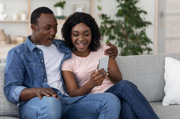 Happy black wife showing her husband photos on smartphone while sitting together on couch in living room. African american smiling couple using mobile phone at home, playing game, copy space
