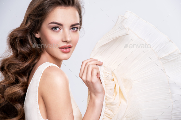 Portrait of a beautiful woman with a long hair in a beige dress. Young brunette model tosses part of her dress – isolated on white. Fashionable girl posing at studio dressed in beautiful light dress