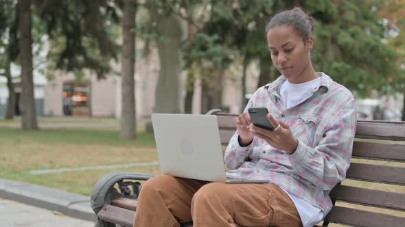 African Woman Using Smartphone and Laptop While Sitting Outdoor on Bench alt