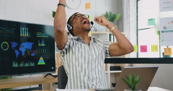 Afro American Man Employee in Headphones Sitting at His Desk and Using Laptop in the Office room alt